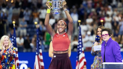 Young Black tennis champ Coco Gauff holds a trophy above her head and grins.