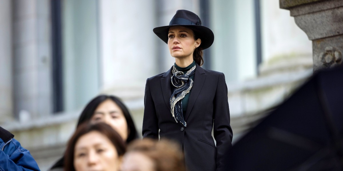 A woman stands in above the crowd wearing black hat and trench coat in 'The Fall of the House of Usher.'