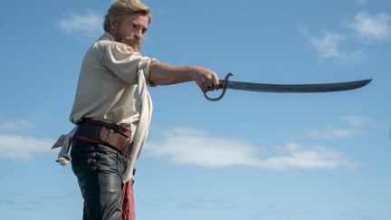 Stede holds a sword out against a backdrop of a blue sky. He has a beard.