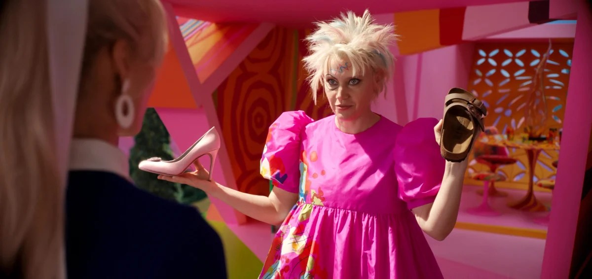 Kate McKinnon as Weird Barbie, with wild hair and pink dress, holding a Birkenstock sandal in one hand and a high heel in the other.