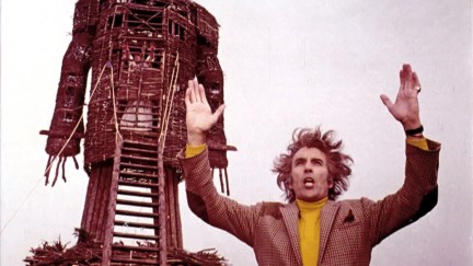 A man gesticulates to a crowd of onlookers while a towering wooden statue stands behind him 