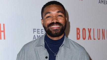 Image of Jordan Howlett on the red carpet at the BoxLunch Holiday Gala 2023. He is a Black man with short black hair and a black beard. He's wearing a grey jacket over a navy blue turtleneck with a silver cross pendant around his neck. He's smiling at the camera and standing in front of a step and repeat that has the BoxLunch and Feeding America logos on it.