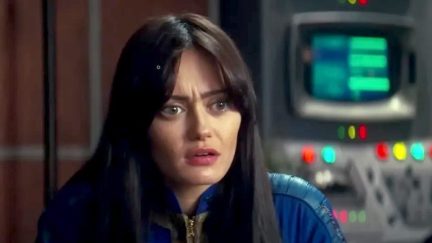 Ella Purnell as Lucy in a scene from Amazon's 'Fallout' series. She is a young white woman with long dark hair and long bangs parted down the middle. She's wearing a blue 'Fallout' vault suit and sitting in a room with computer consoles behind her. She has a confused expression on her face.