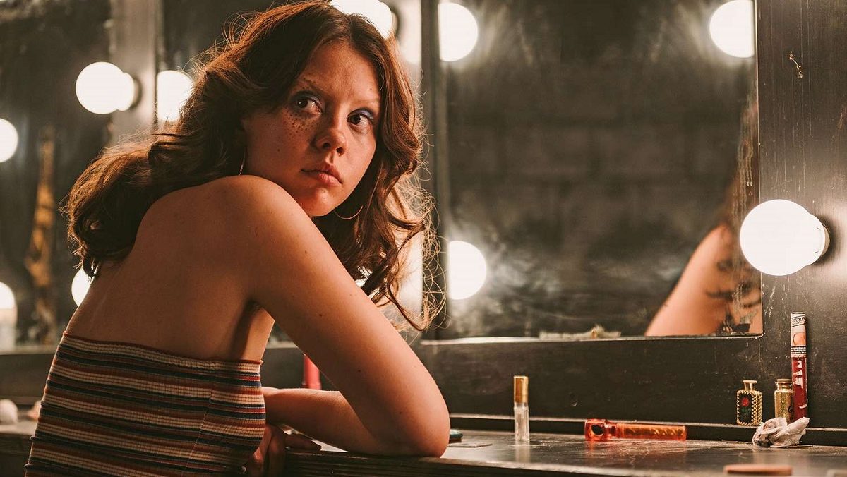 Mia Goth in a scene from the film 'X.' She is a young white woman seated at a make-up table with large bulbs around the mirror. Her arms are folded on the table as she looks over her shoulder. She has long, brown hair and is wearing a red, black, and white striped tube top. It's a 1970s period look.