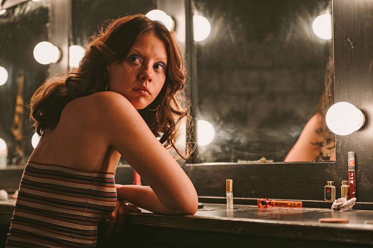 Mia Goth in a scene from the film 'X.' She is a young white woman seated at a make-up table with large bulbs around the mirror. Her arms are folded on the table as she looks over her shoulder. She has long, brown hair and is wearing a red, black, and white striped tube top. It's a 1970s period look.