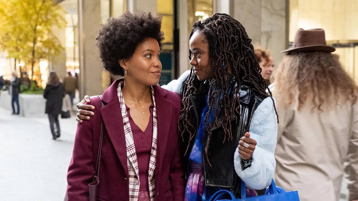 Image of Sinclair Daniel and Ashleigh Murray in a scene from 'The Other Black Girl.' They are both young Black women walking down a city street. Daniel is wearing her hair in a medium afro and wearing a maroon coat over a maroon and white plaid blazer and maroon v-neck sweater. Murray has long black hair in tight braids. She's wearing a coat with light blue furry sleeves and a black leather torso over a blue and purple print dress. Murray has her arm around Daniel as they look at each other.