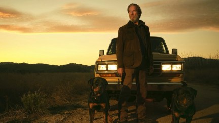 An old man stands in the desert in front of a truck with two dogs in 