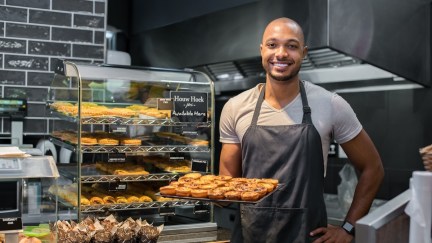 Smiling Black baker with apron holding tray of small pastry and looking at camera.