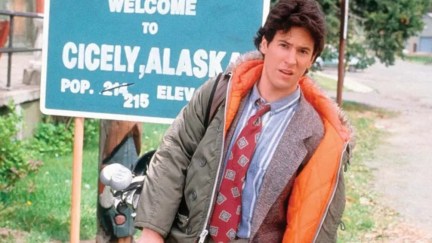 Rob Morrow in Northern Exposure; image depicts a white man with brown hair in a winter coat and suit, standing in from of sign for Cecily, Alaska