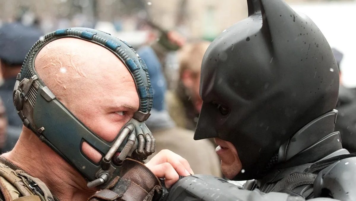 Tom Hardy as Bane and Christian Bale as Batman in a scene from 'The Dark Knight Rises.' Bane is a white man who is bald and wearing a metal mask clamped over the top of his head, and over his cheeks, nose and mouth. Batman is wearing his signature cowl over his head, revealing only his mouth. they are grappling mid-fight.