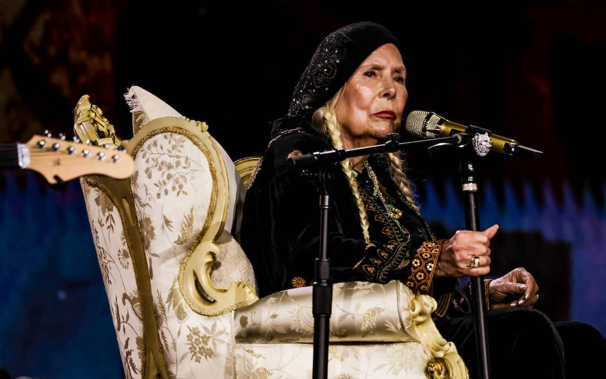 Joni Mitchell sitting on a chair with a microphone in front of her, singing at the Grammys