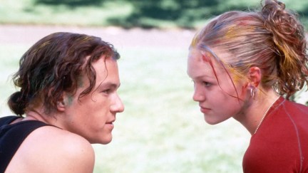 Image of Heath Ledger as Patrick and Julia Stiles as Kat in a scene from '10 Things I Hate About You.' They are both white teenagers sitting on steps staring into each others' eyes. Patrick has wavy, chin-length brown hair and is wearing a black tank top. His hair's pulled back into a short pony tail and there is green paint in his hairline. Kat has wavy blonde hair pulled up into a ponytail, and has various colors of paint in her hair. She's wearing a red shirt.