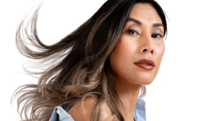 Headshot of Rain Valdez. She is a brown Asian trans woman with long dark hair with blonde highlights blowing in the breeze. Her body is turned to the side with her face turned to the camera. She's wearing a light blue buttondown that's open at the top and coming off her shoulder.