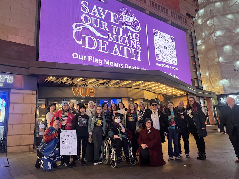 A crowd poses for a photo underneath a purple billboard that reads "Save Our Flag Means Death."