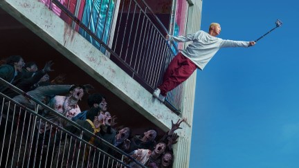 A boy hangs out the window of an apartment complex filming zombies with a selfie stick in 