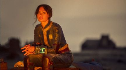 Image of Ella Purnell as Lucy in Prime Video's 'Fallout.' She is a white woman with long, dark hair pulled into a ponytail and wearing a blue 'Fallout' vault suit. She has a Pip-Boy on her wrist and she's sitting on a box in front of a fire at night, resting her arms on her lap as she looks out into the distance.