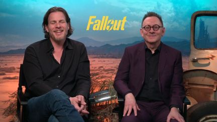 Image of Jonathan Nolan and Michael Emerson sitting in director chairs during a virtual press junket. Nolan is a white man with chin-length wavy brown hair and a thin beard wearing a black buttondown and jeans. Emerson is an older white man with closely cropped black hair that sticks straight up wearing black-rimmed glasses and a maroon suit with a black buttondown shirt.