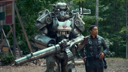 Image of Knight Titus and Maximus (played by Aaron Moten) in a scene from Prime Video's 'Fallout.' Knight Titus is in a suit of silver T-60 power armor with a red Brotherhood of Steel insignia on the chest. He's holding an enormous gun. Maximus is a young Black man with close cropped dark hair wearing a Brotherhood uniform under a parachute harness. They are walking through a wooded area.