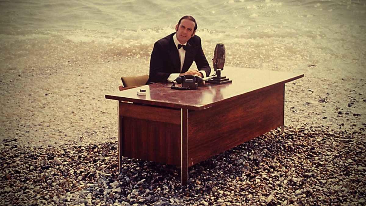John Cleese sits at a desk on the beach