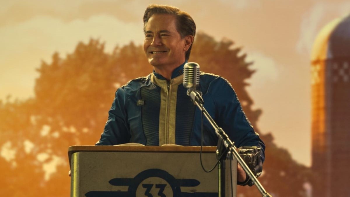 Kyle MacLachlan as Hank MacLean in a scene from Prime Video's 'Fallout.' He is a white man with short dark hair parted on the side wearing a blue and yellow vault suit and standing at a podium with the Vault 33 logo on it. Behind him is a projected image of a sunny farm.