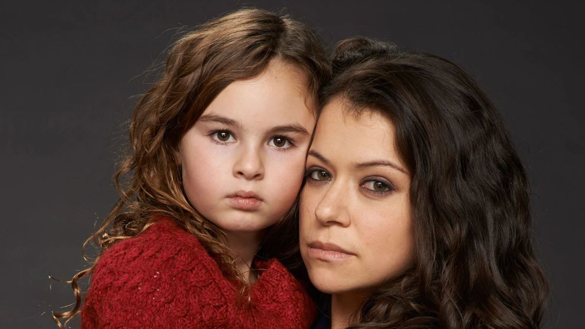 Skyler Wexler as Kira and Tatiana Maslany as Sarah in a promotional photo for BBC America's 'Orphan Black.' Kira is a white young girl with long, wavy brown hair wearing a red sweater. She is being carried by Sarah, a white woman with long, wavy dark brown hair. They both have brown eyes.