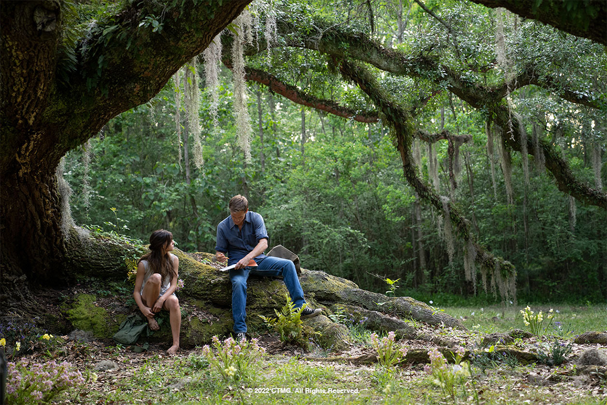 A teen girl and boy sit beneath a massive tree