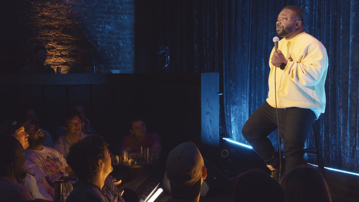 Brian Simpson sits on a chair on stage while audience laughs