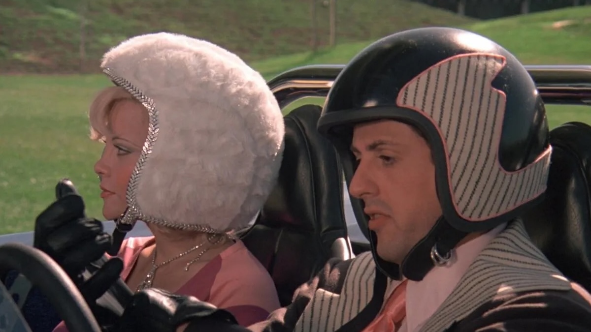 Sylvester Stallone is driving a convertible wearing a helmet and has a woman next to him.
