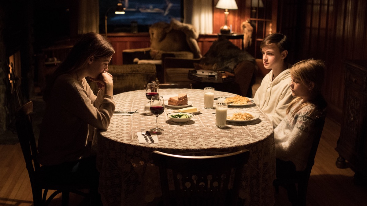 A woman sits at a dinner table in a cabin with two kids in "The Lodge" 