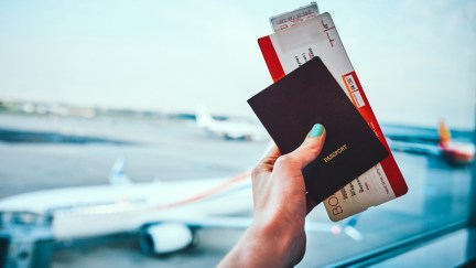 A hand holds a passport and other documents in front of an airplane