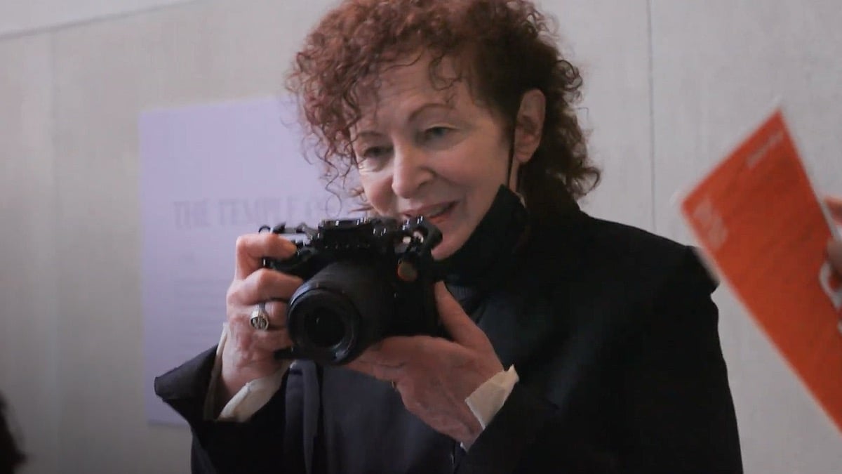 A woman in a mask holds a camera in "All the beauty and the Bloodshed"