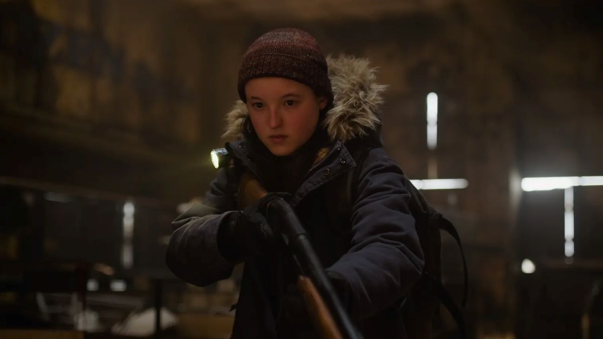 Bella Ramsey as Ellie in a scene from season 2 of HBO's 'The Last of Us.' Ellie is a young, white woman wearing a dark red winter hat and a black parka with grey fur around the hood. She's standing in a barn and holding a rifle.