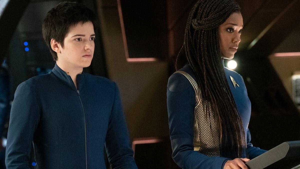 Blu del Barrio as Adira Tal and Sonequa Martin-Green as Michael Burnham in a scene from 'Star Trek: Discovery.' Del Barrio is a white, non-binary Latine with short dark hair wearing a dark blue zip-up jumpsuit. Martin-Green is a Black woman with long, black braids wearing a dark blue and gold Starfleet uniform. She is standing at a console while del Barrio stands behind her.