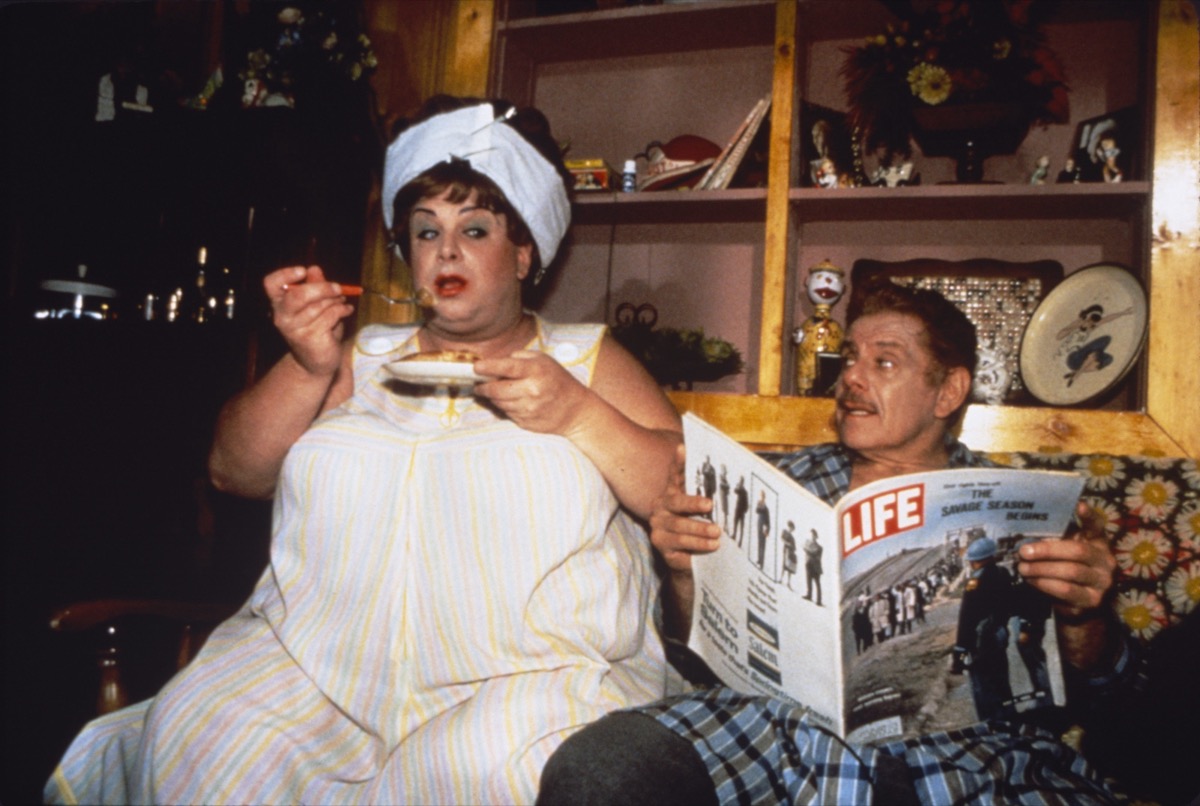 A man and a woman sit on a couch in "Hairspray", the man reads the paper while the woman eats. 