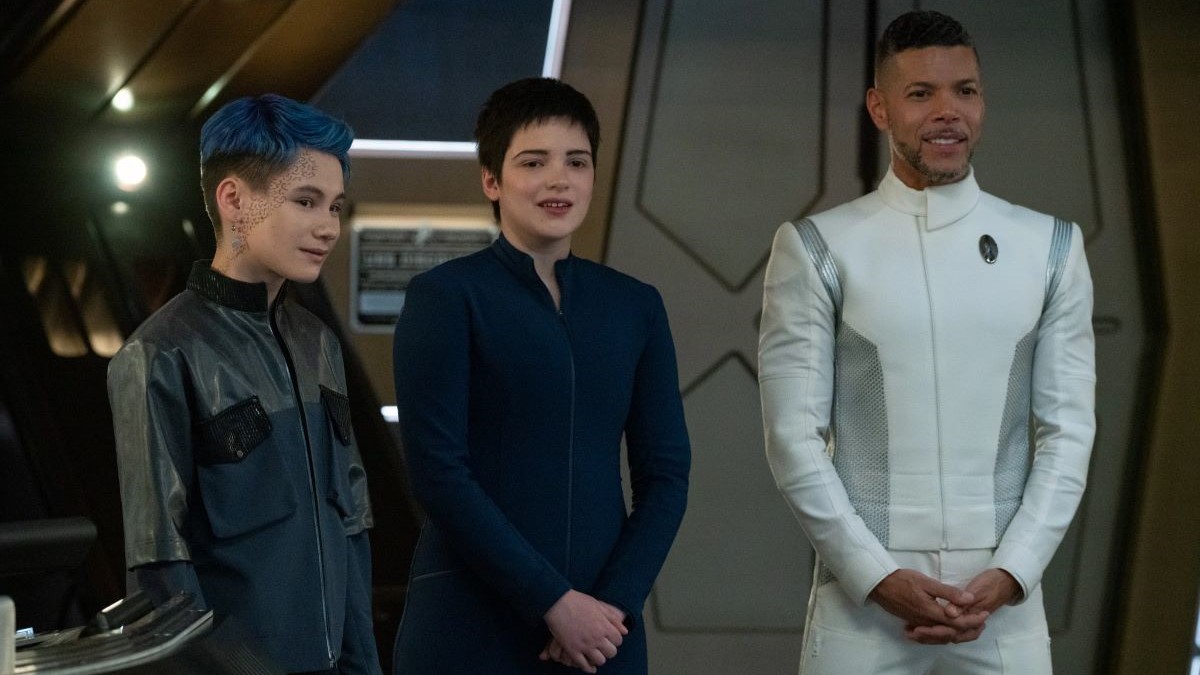 Ian Alexander as Grey, Blu Del Barrio as Adira, and Wilson Cruz as Culber in a scene from 'Star Trek: Discovery." Alexander is a Vietnamese trans masc person with short blue hair and wearing a long black jacket. Del Barrio is a white Latine non-binary person with short dark hair wearing a blue Starfleet uniform. Cruz is an older Latino man with short, dark hair and a light salt-and-pepper beard wearing a white Starfleet uniform. They are standing in a row and all looking at the same thing, smiling.