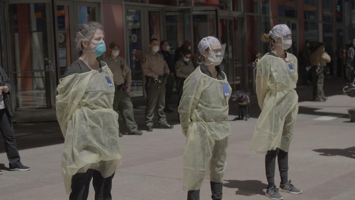 Three medical personnel stand on the street wearing PPE in "In The Same Breath"