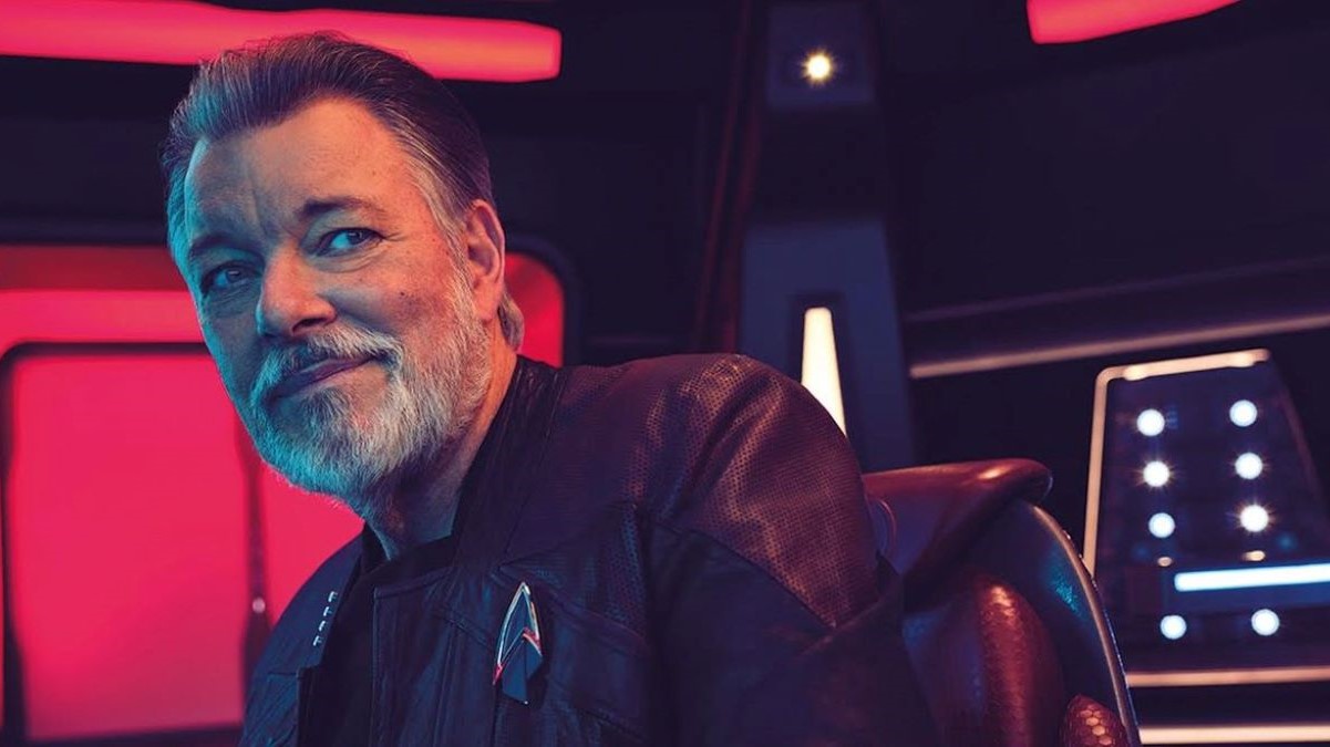 Image of Jonathan Frakes as Will Riker on 'Star Trek: Picard.' He is a white man with dark hair and a grey beard wearing a black Starfleet uniform. He's seated in a captain's chair and glancing to the side, smirking.