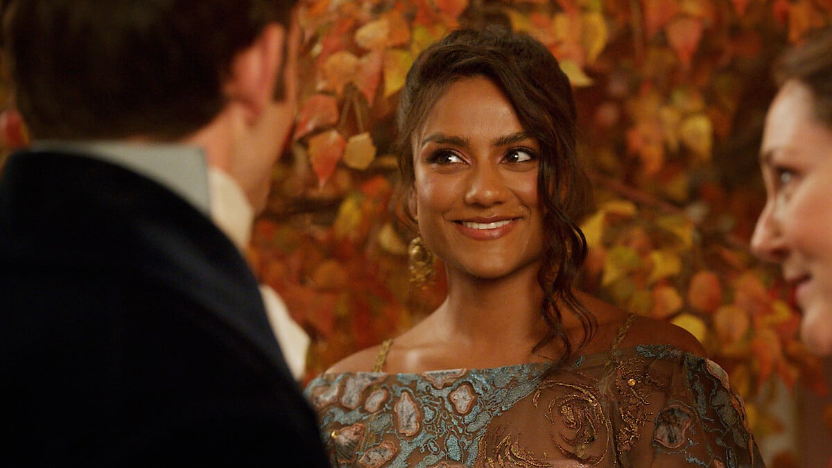 Image of Simone Ashley as Kate in a scene from Netflix's 'Bridgerton.' She is an Indian woman with long, dark hair pulled up into a Regency-era hairstyle with wavy tendrils hanging down one side of her face. She is smiling and wearing a light blue and gold-patterned dress.