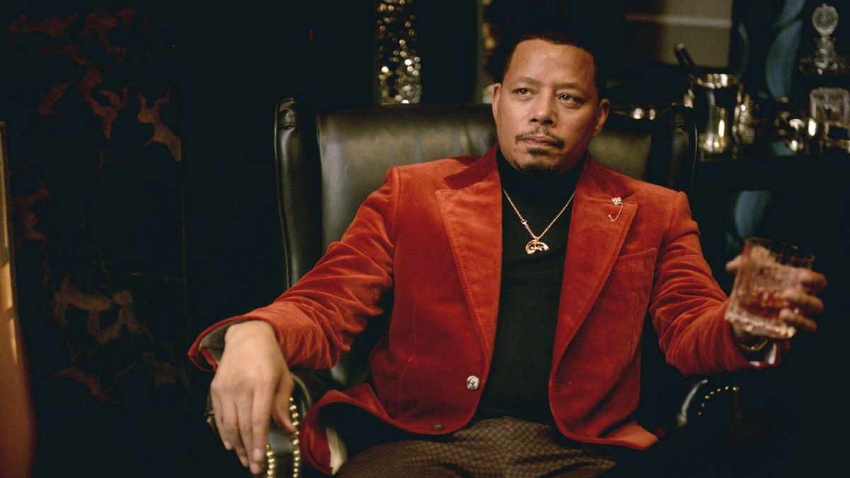 Image of Terrence Howard as Lucious Lyon in a scene from 'Empire' on FOX. He is a Black man with close-cropped dark hair and a thin goatee. He's wearing a red velvet blazer over a black turtleneck with a thin gold chain and brown pants. He's seated in a black armchair and holding a glass of scotch.