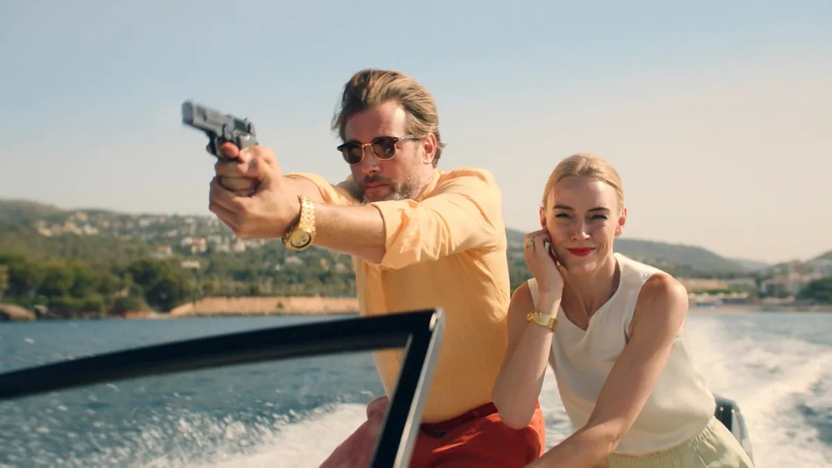 Julian Looman as Max Winter, holding up a gun on a speedboat, with Elen Rhys as Miranda Blake sat in the background. 