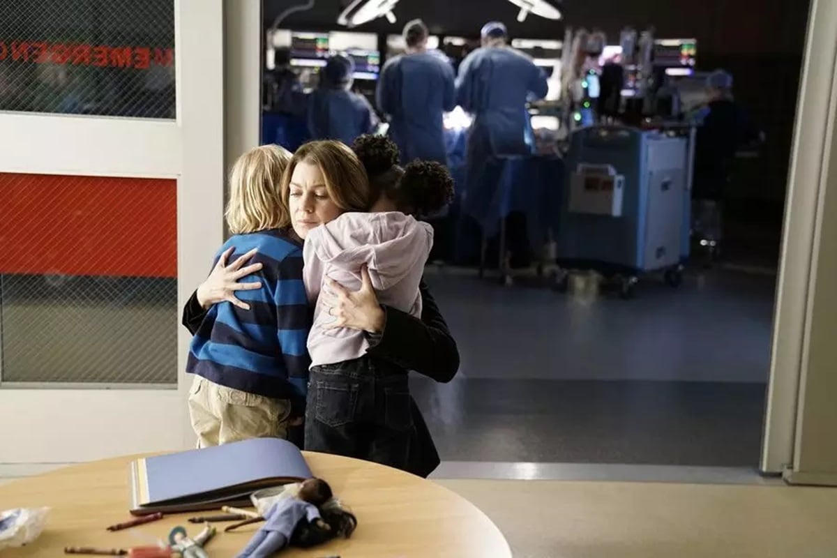 Meredith hugs two children while surgeons operate on a patient in the background