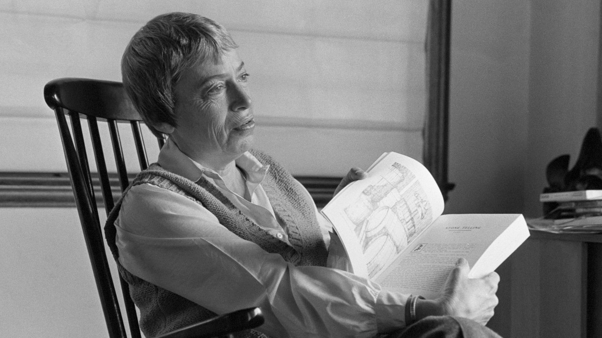 Ursula K. Le Guin'sitting in a rocking chair and holding a book, black and white photograph.