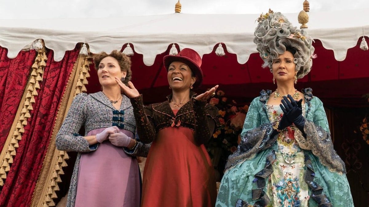 Ruth Gemmell as Violet, Adjoa Andoh as Lady Danbury, and Golda Rosheuvel as Queen Charlotte on Netflix's 'Bridgerton.' They are standing in a row looking out off a stage and cheering something. Violet is a white, middle-aged woman with brown hair in a purple and grey dress. Lady Danbury is an older Black woman in a red and black dress and a red hat. Queen Charlotte is a middle-aged Black woman in a tall, grey wig wearing an elaborate blue gown.