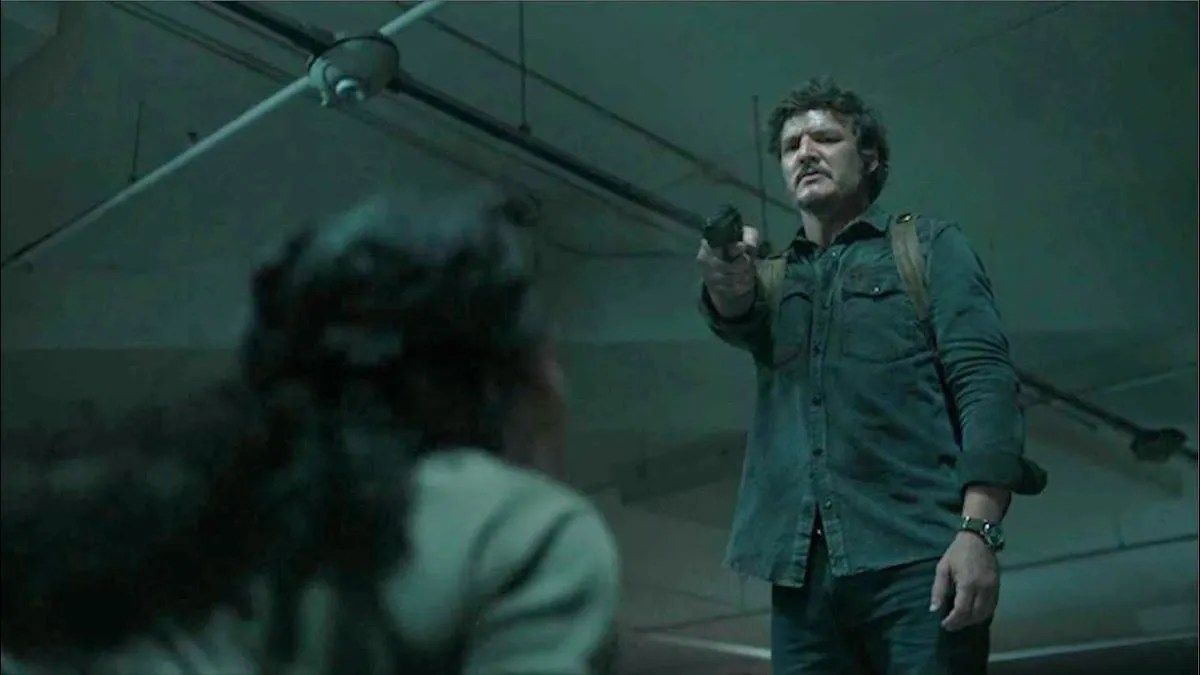 Pedro Pascal as Joel in a scene from HBO's 'The Last of Us." He is a white Latino man with shaggy dark hair, a mustache and facial stubble. He's wearing a dark green button-up, jeans, and a brown backpack and he's holding a gun on Merle Dandridge's Marlene, who is on the floor with her back turned to the camera.