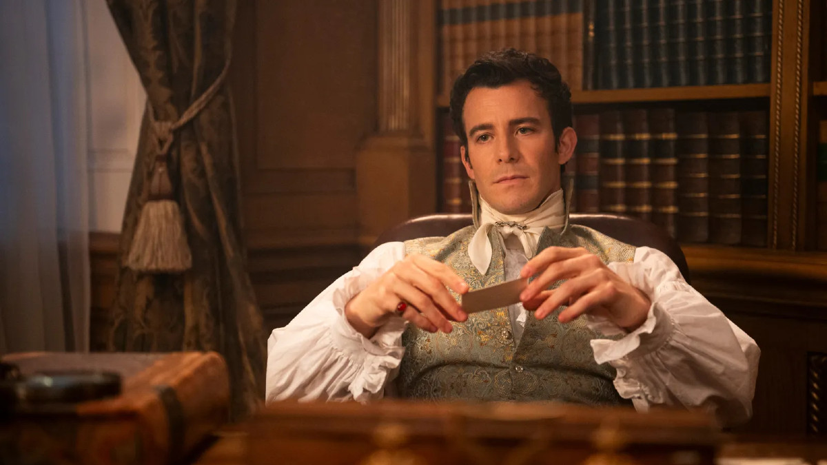 Luke Thompson as Benedict Bridgerton in Netflix's Bridgerton, sitting at a desk and holding a card in his hands.
