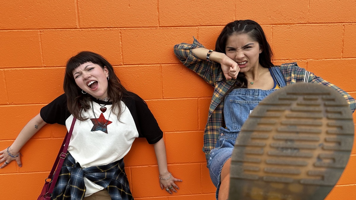 Two teen girls stand against an orange wall. One kicks her foot so we see the bottom of her shoe