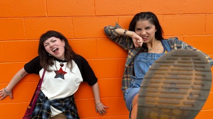 Two teen girls stand against an orange wall. One kicks her foot so we see the bottom of her shoe