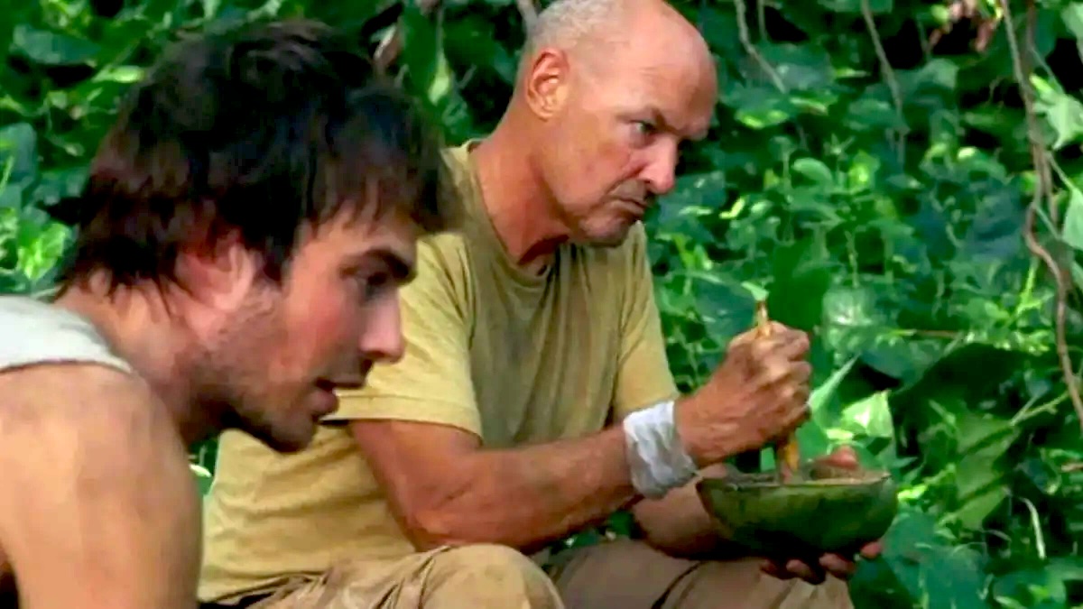 Ian Somerhalder as Boone and Terry O'Quinn as Locke in a scene from ABC's 'Lost.' They are sitting at a campsite in a jungle surrounded by green foliage. Boone is a young white man with dark shaggy hair wearing a grey tank top. Locke is an older, bald white man wearing a light green t-shirt and khaki pants. He's using a mortar and pestle to ground something down.