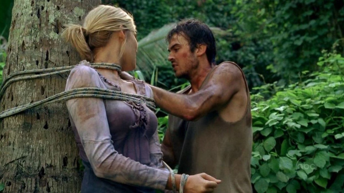 Maggie Grace as Shannon and Ian Somerhalder as Boone in a scene from ABC's 'Lost.' Shannon is a young, white woman with her blonde hair in a ponytail. She is tied to a tree and wearing a long-sleeved purple blouse. Boone is a young, white man with shaggy dark hair wearing a grey tank top. He is cutting her loose from the tree.