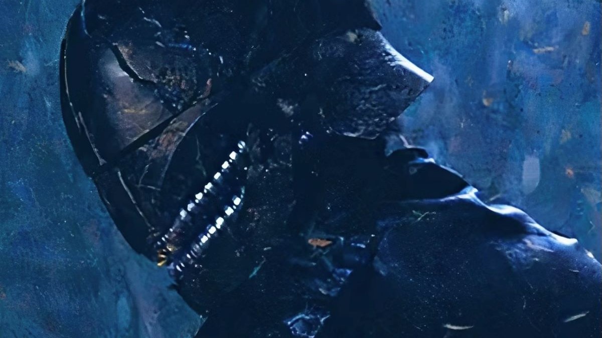 Manny Jacinto as Qimir in the Disney+ Star Wars show, 'The Acolyte.' It's a close-up of Qimir turned to the side in a black helmet that covers his whole face. It has silver detailing around the mouth area that make it look like the helmet has a big creepy smile. He's dressed all in black.